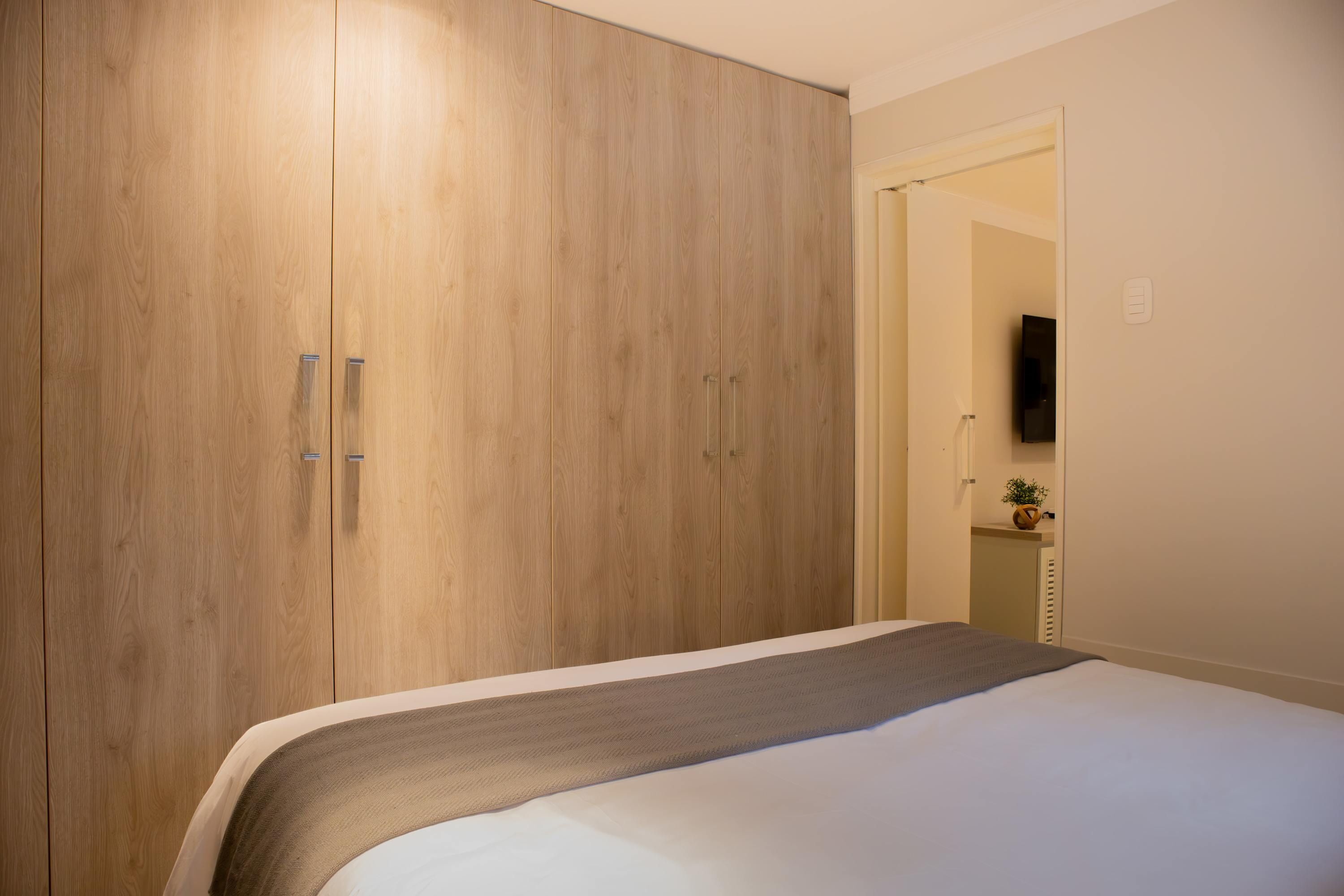 Een slaapkamer met een grote kast, een bed met een grijze deken, een flatscreen televisie aan de muur en een kleine plant op een plank.