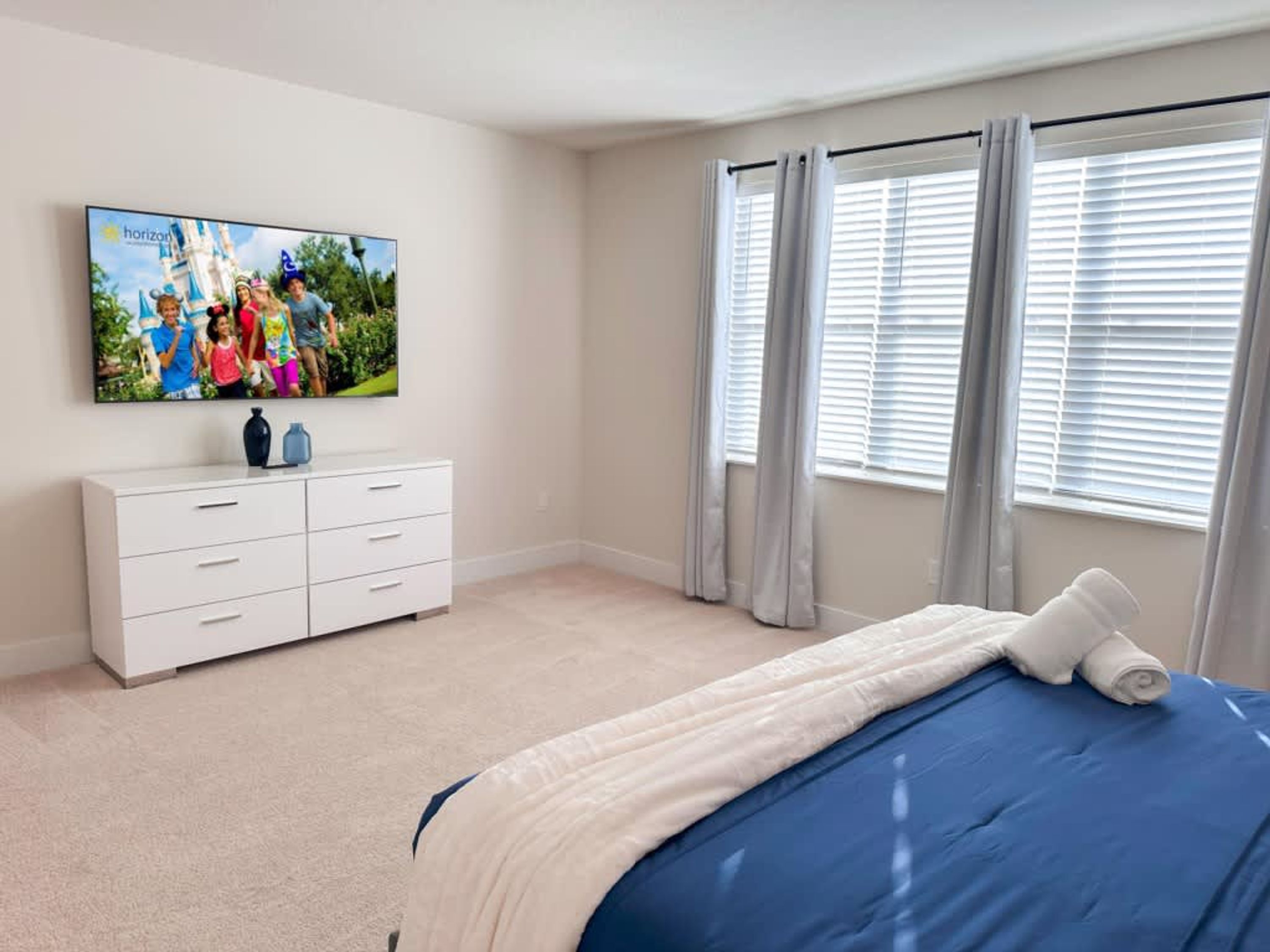 A bedroom with a bed, a dresser, a television on the wall, and a window with curtains.