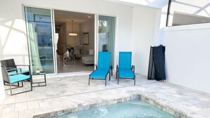 A swimming pool with two blue chairs and a table, a glass door leading to a living area, and a white wall.
