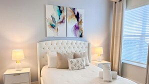 A neatly arranged bedroom with a tufted headboard, two bedside tables with lamps, a bed with white bedding, and abstract wall art.