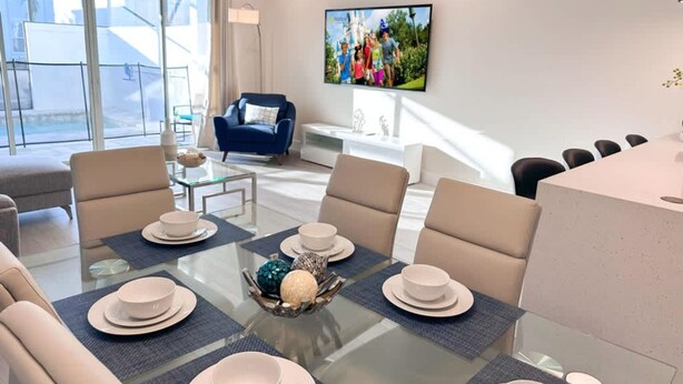 A modern living room with a glass dining table set for six, a blue armchair, and a television mounted on the wall.