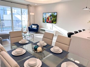 A modern living room with a glass dining table set for six, a blue armchair, and a television mounted on the wall.
