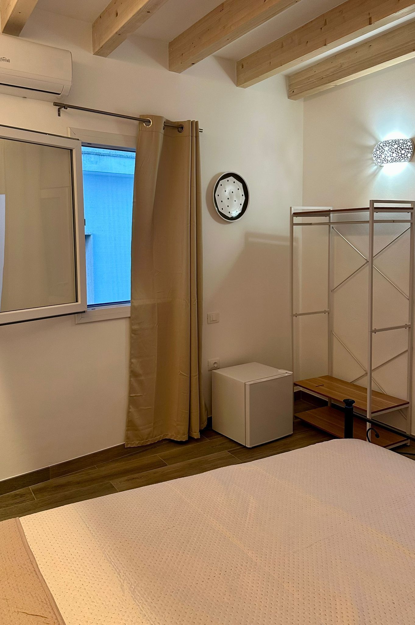 A bedroom with a bed, a small bedside table, a wall-mounted heater, a window with curtains, and a wooden ceiling beam.