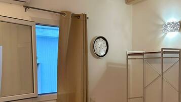 A bedroom with a bed, a small bedside table, a wall-mounted heater, a window with curtains, and a wooden ceiling beam.