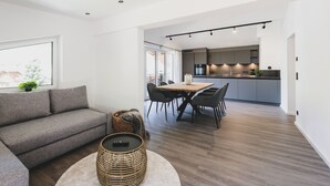 Een moderne woonkamer met een grijze bank, een ronde rieten salontafel, en een eethoek met een houten tafel en stoelen.