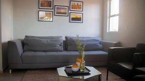 Ein modernes Wohnzimmer mit einem grauen Sofa, einem Glastisch und einem schwarzen Sessel. An der Wand hängen Bilderrahmen und rechts befindet sich ein Fenster.