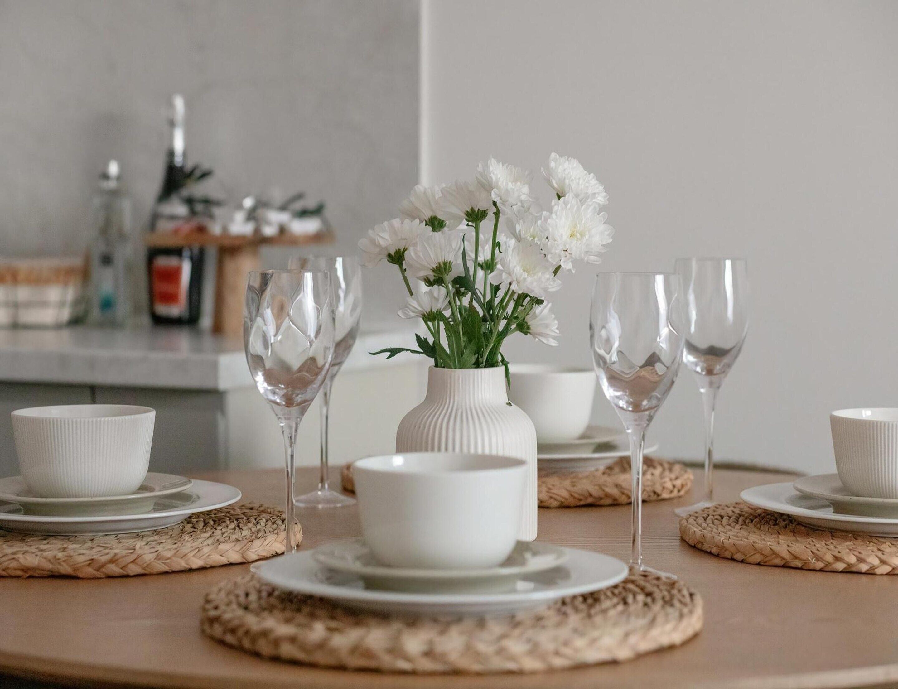 Ett bord dukat med vita tallrikar, skålar och vinglas, med en vit vas med blommor som mittpunkt.