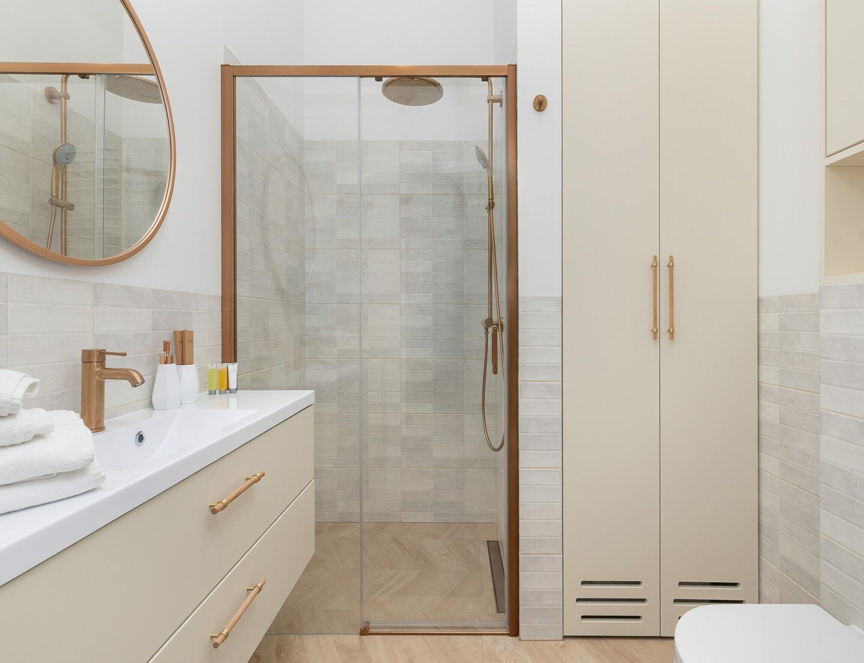 Un baño moderno con ducha envuelta en vidrio, un lavamanos blanco con detalles dorados y un mueble beige con espejo.