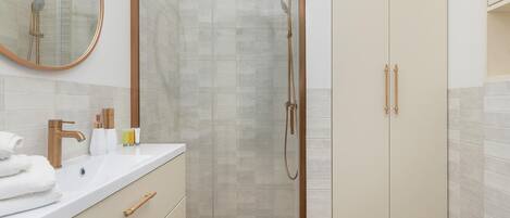 Ein modernes Badezimmer mit einer gläsernen Dusche, einem weißen Waschbecken mit goldfarbenen Armaturen und einem beige gefärbten Schrank mit Spiegel.