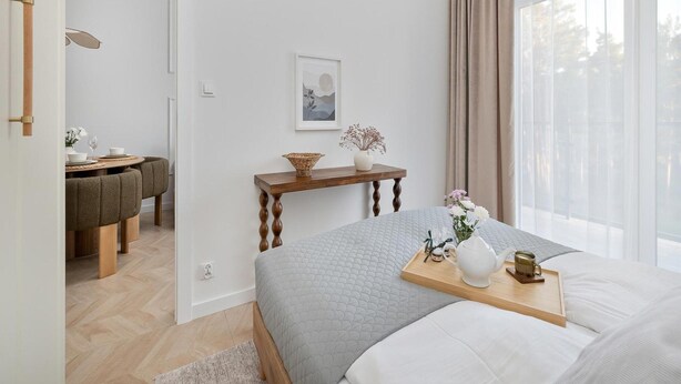 A modern bedroom with a large bed, a bedside table, a small table with a vase, and a view of the outdoors through large windows. - Apartment, Indoor Pool, Close to the Beach (Rewal)