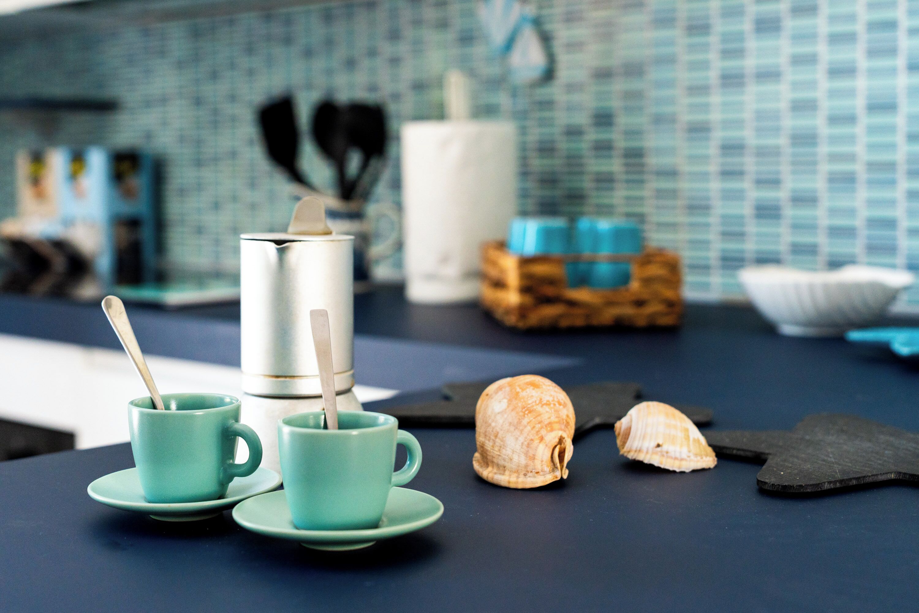 A kitchen counter with two teal cups and saucers, a white kettle, a seashell, and a black spatula.