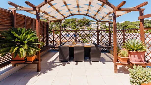 Un espacio de estar al aire libre cubierto con una mesa y sillas, rodeado de plantas en macetas y una pérgola de madera.