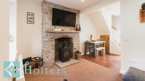 A living room with a stone fireplace, a flat-screen TV mounted above it, and a dining area with a wooden table and chairs.