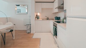 Una cocina moderna con armarios blancos, horno empotrado y lavaplatos. Hay una mesa de comedor con sillas y un cubo de basura.