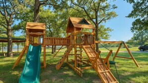 A wooden playground structure with a slide and climbing elements, surrounded by trees and grass.
