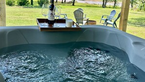 A hot tub with a wooden tray holding wine glasses and a bottle, surrounded by outdoor chairs and a view of a grassy area with trees.