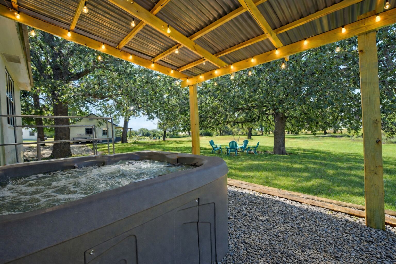 A hot tub under a wooden pergola with string lights, surrounded by a grassy area with trees and outdoor seating.