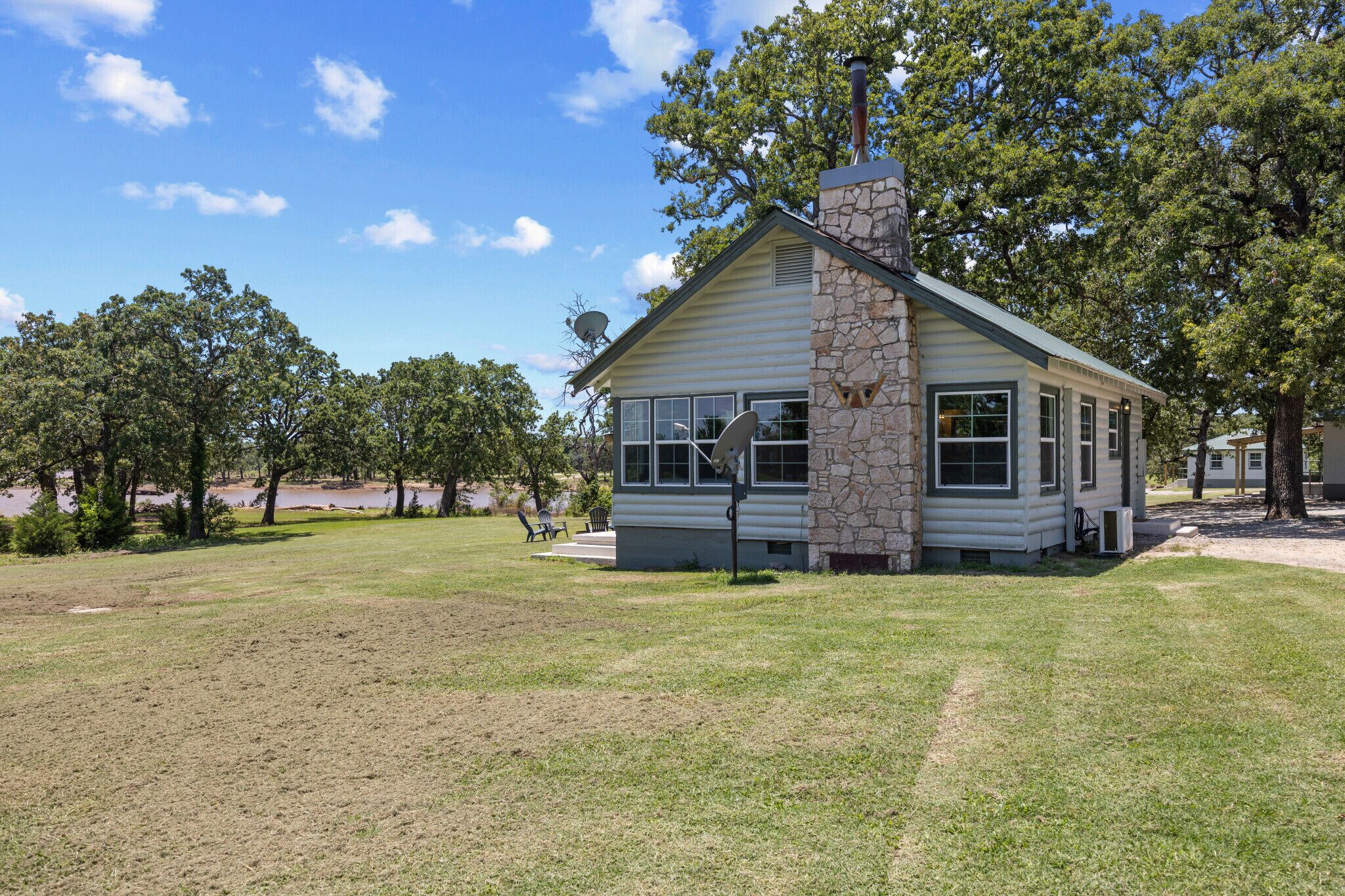 A small, single-story log cabin with a stone chimney, surrounded by a grassy lawn and trees.