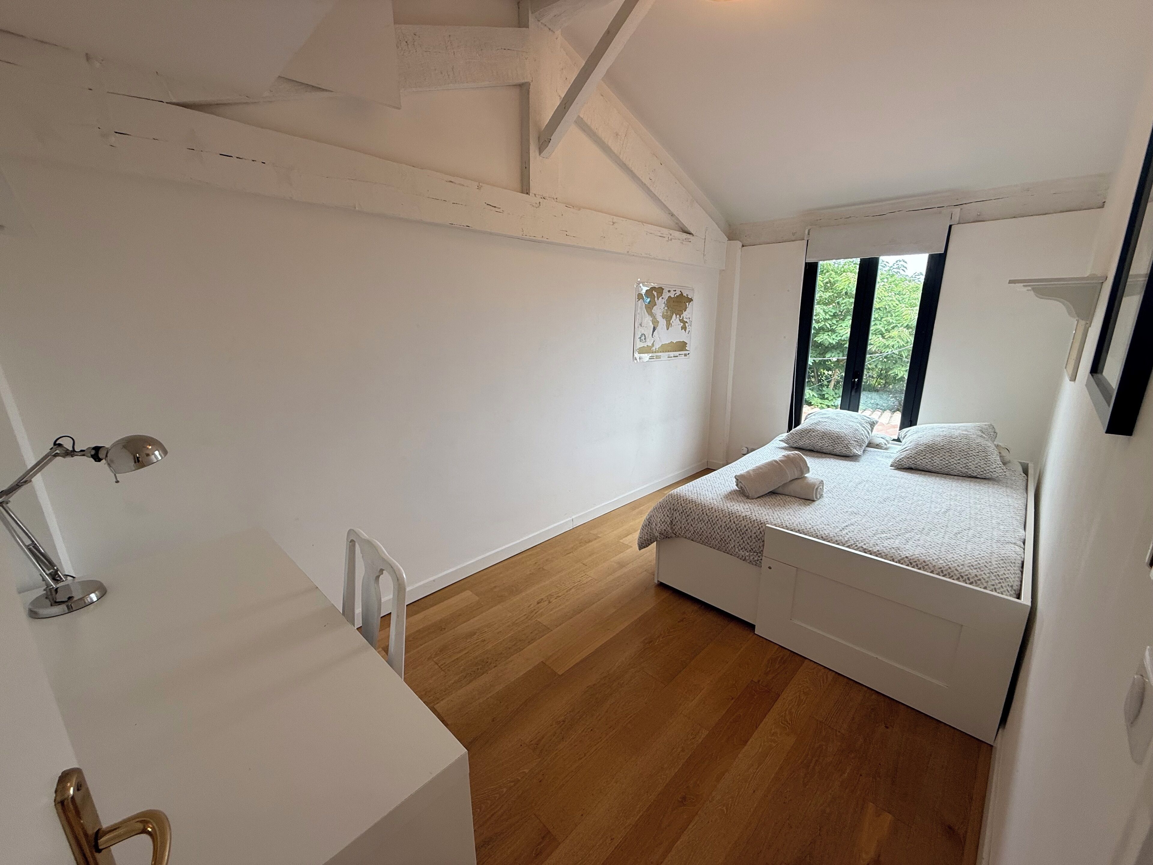 A bedroom with a bed, a desk, and a chair. There is a window with a view of greenery outside.