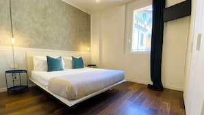 Un dormitorio moderno con una cama grande, dos mesitas de noche, una lámpara de pared y una ventana con vistas a edificios.