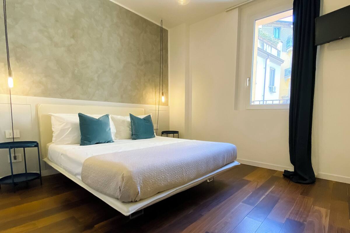 A modern bedroom with a large bed, two bedside tables, a wall-mounted light, and a window with a view of buildings.