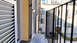 Un balcón estrecho con barandillas de metal, suelo de baldosas y vistas a edificios vecinos.