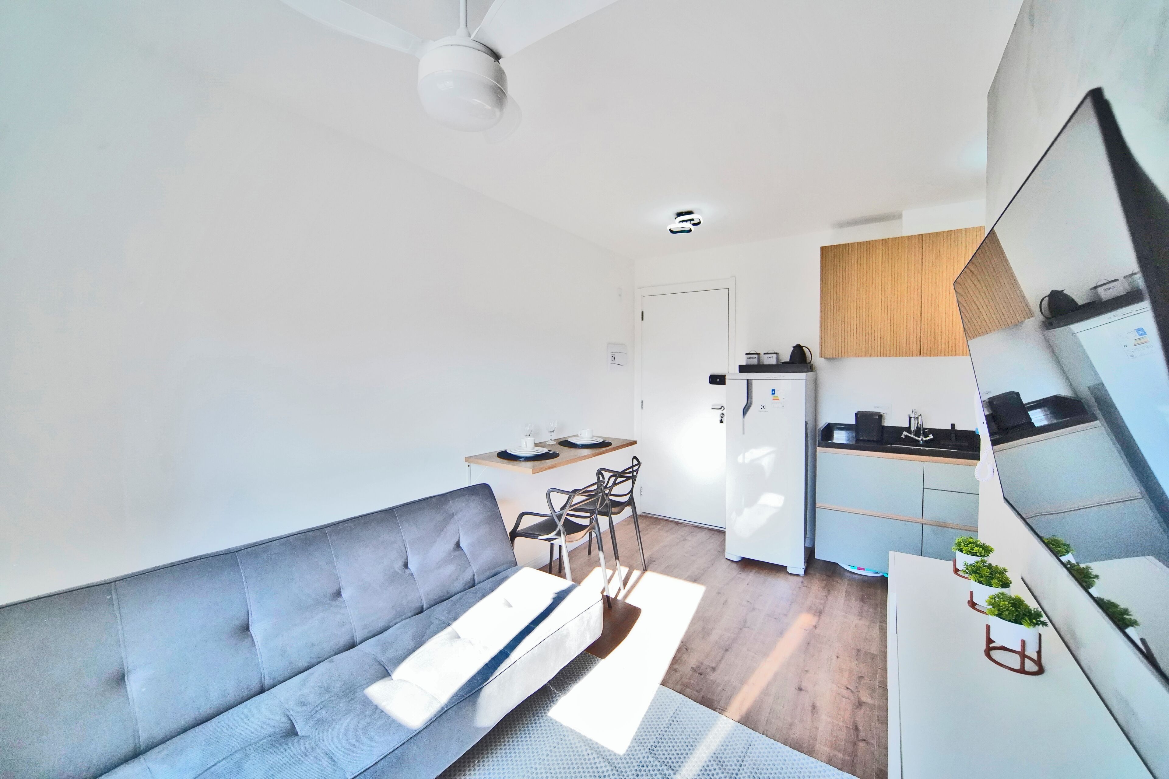 Un espacio habitable moderno y funcional con un sofá gris, una zona de comedor con sillas, una cocina con refrigerador y un ventilador de techo.
