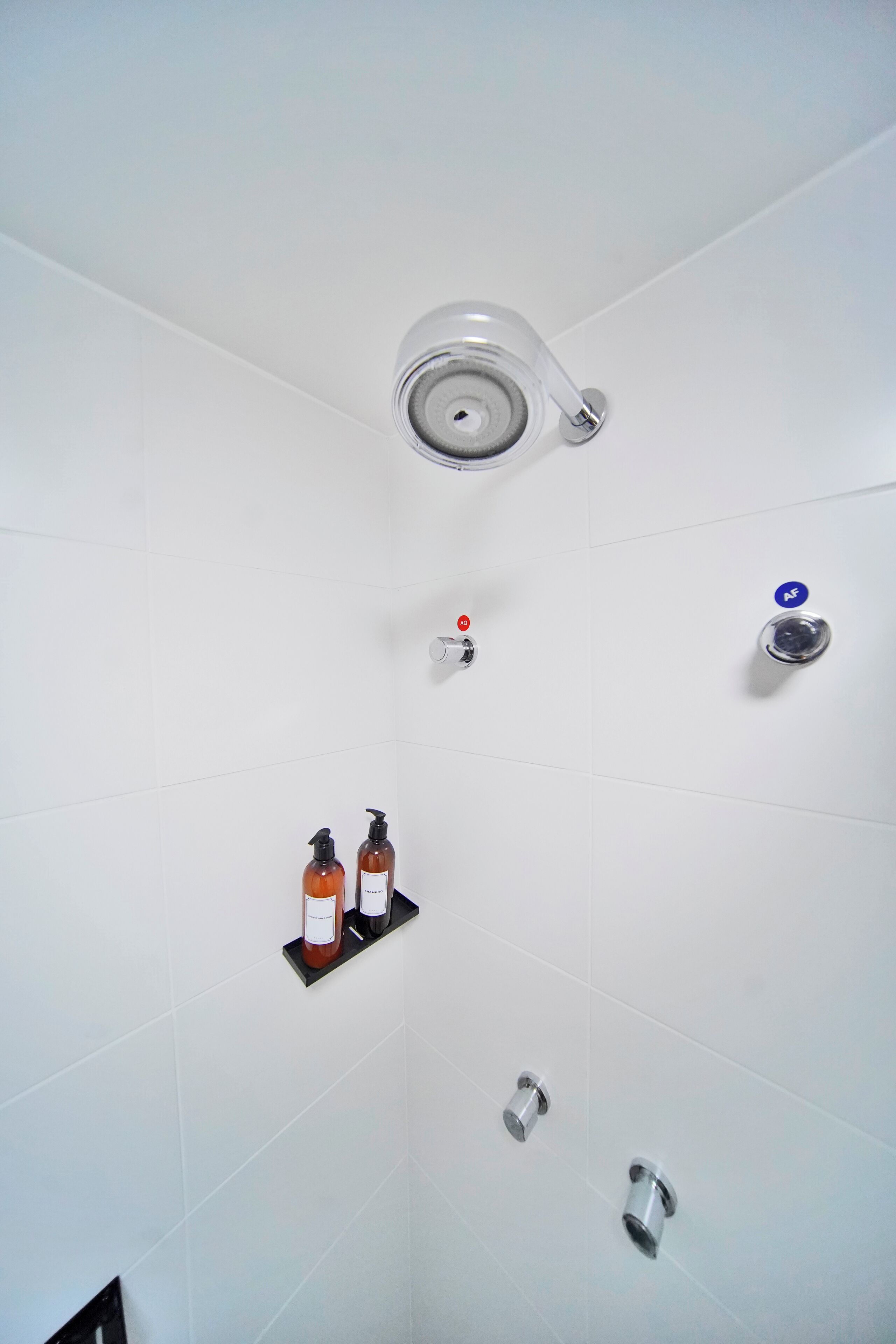Un baño moderno con azulejos blancos, cabezal de ducha, grifo y dos dispensadores de jabón en una estantería.