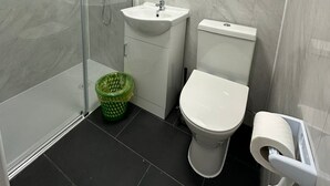 A bathroom with a glass-enclosed shower, a sink, a toilet, and a green trash bin. - Comfortable Budget Ensuite Room in East London 3 (London)
