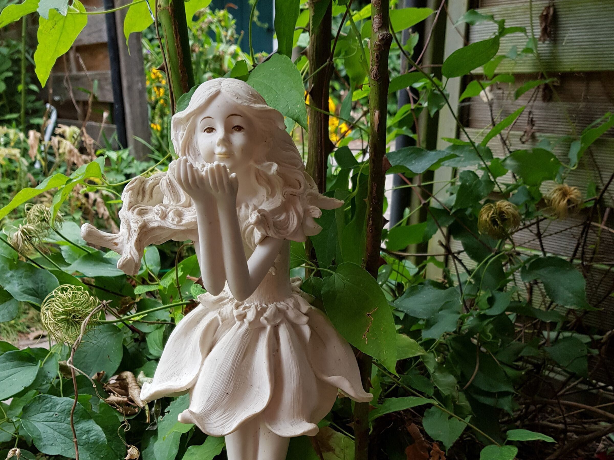 En statue af en pige i kjole, omgivet af grøn vegetation.
