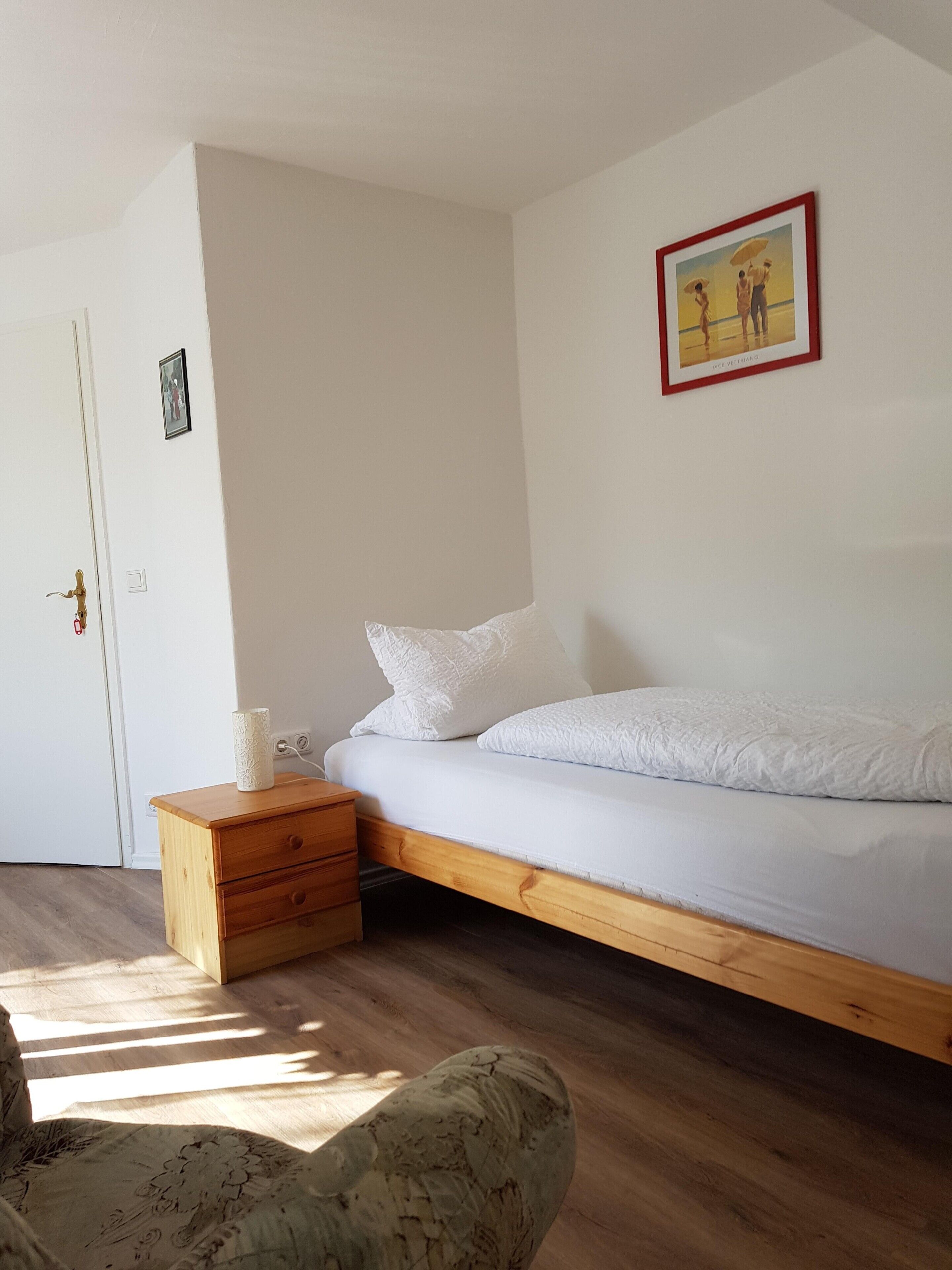 Une chambre à coucher avec un lit, une table de chevet en bois, une petite table, une porte et un tableau encadré au mur.