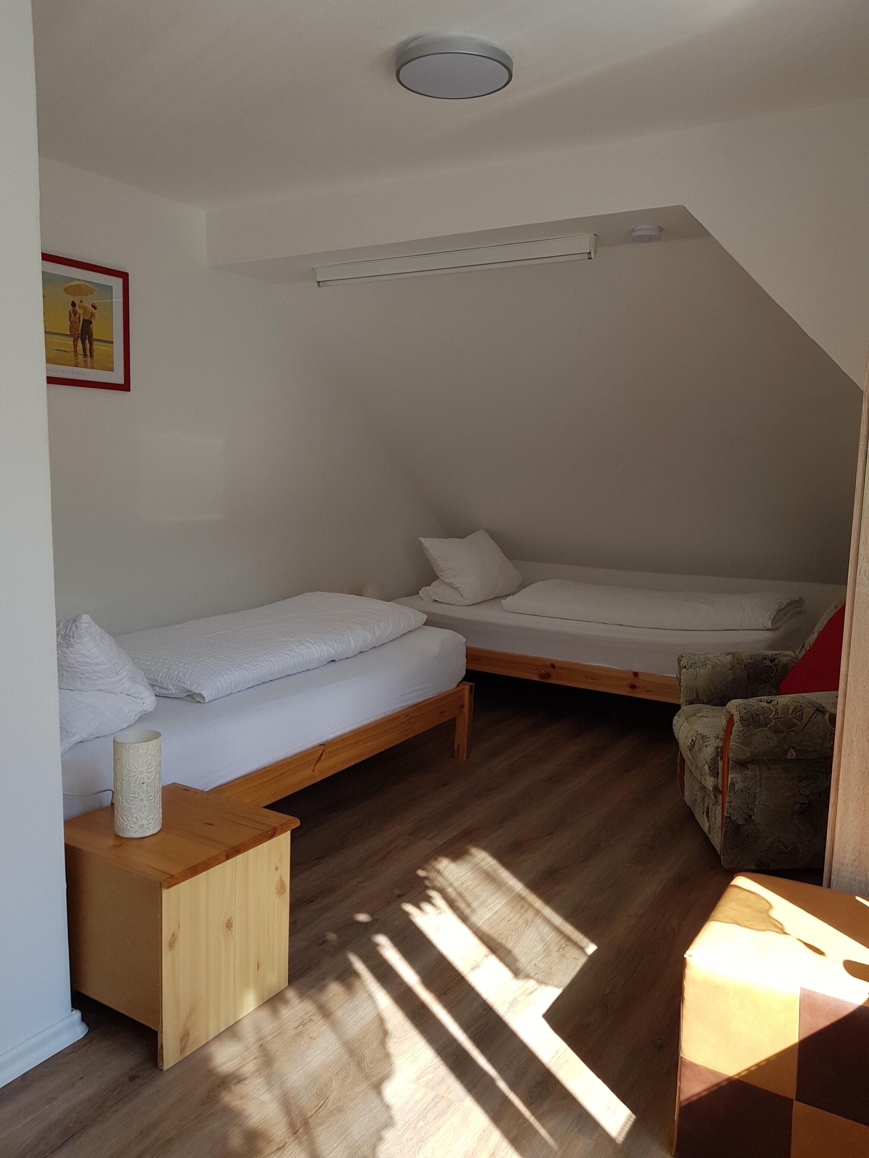 A small, well-lit room with two beds, a wooden nightstand, a patterned armchair, and a ceiling light fixture.