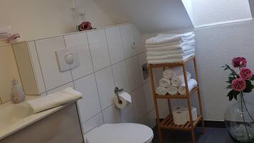 Une salle de bain comprenant des toilettes, un porte-serviettes, une étagère avec des serviettes, un support à papier toilette et un puits de lumière.