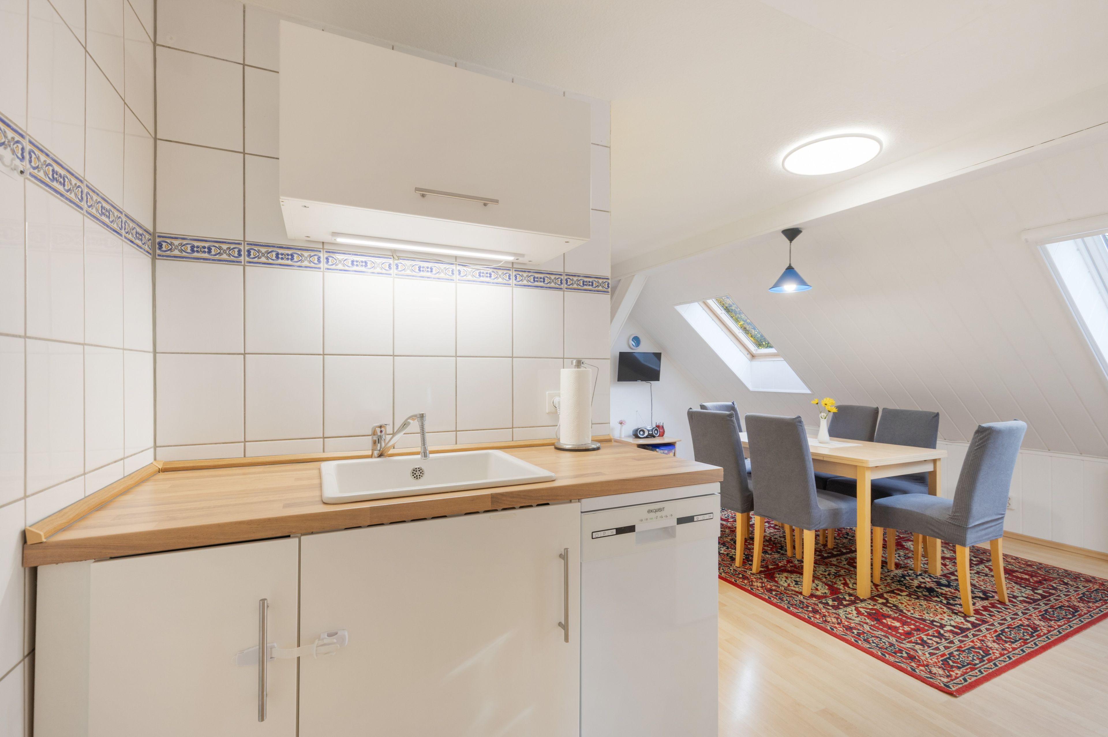 Een kleine keuken met een spoelbak, een houten aanrechtblad en een eethoek met een tafel en stoelen.