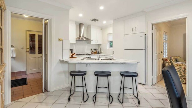 A modern kitchen with white cabinets, a stainless steel range hood, a central island with stools, and a refrigerator. - Maison d'Art (Glenside)