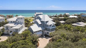 A large, multi-story house with a complex roof design, surrounded by lush greenery and a view of the ocean.