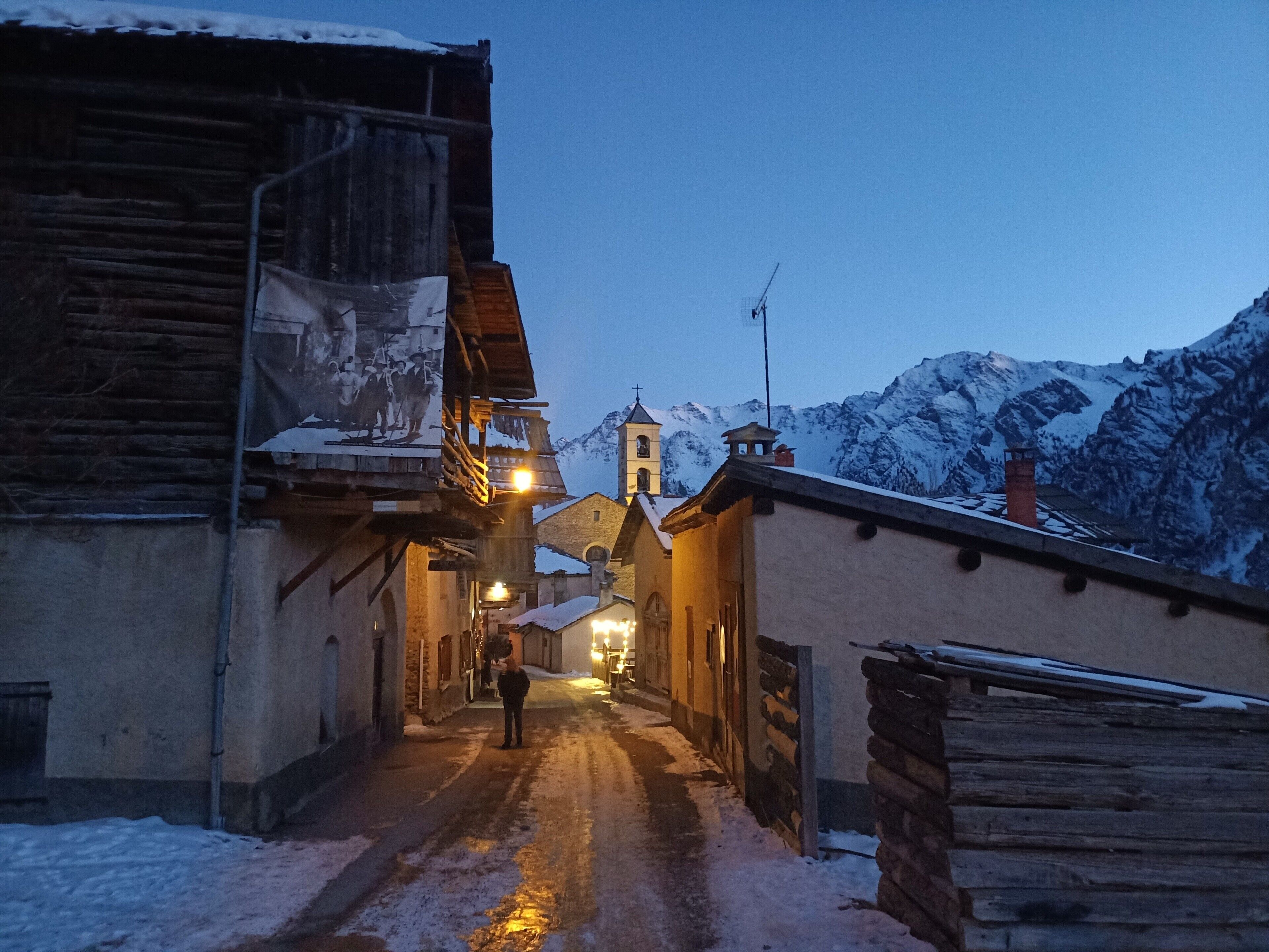 Una calle estrecha cubierta de nieve con edificios a ambos lados, una torre de campanas y montañas nevadas al fondo.