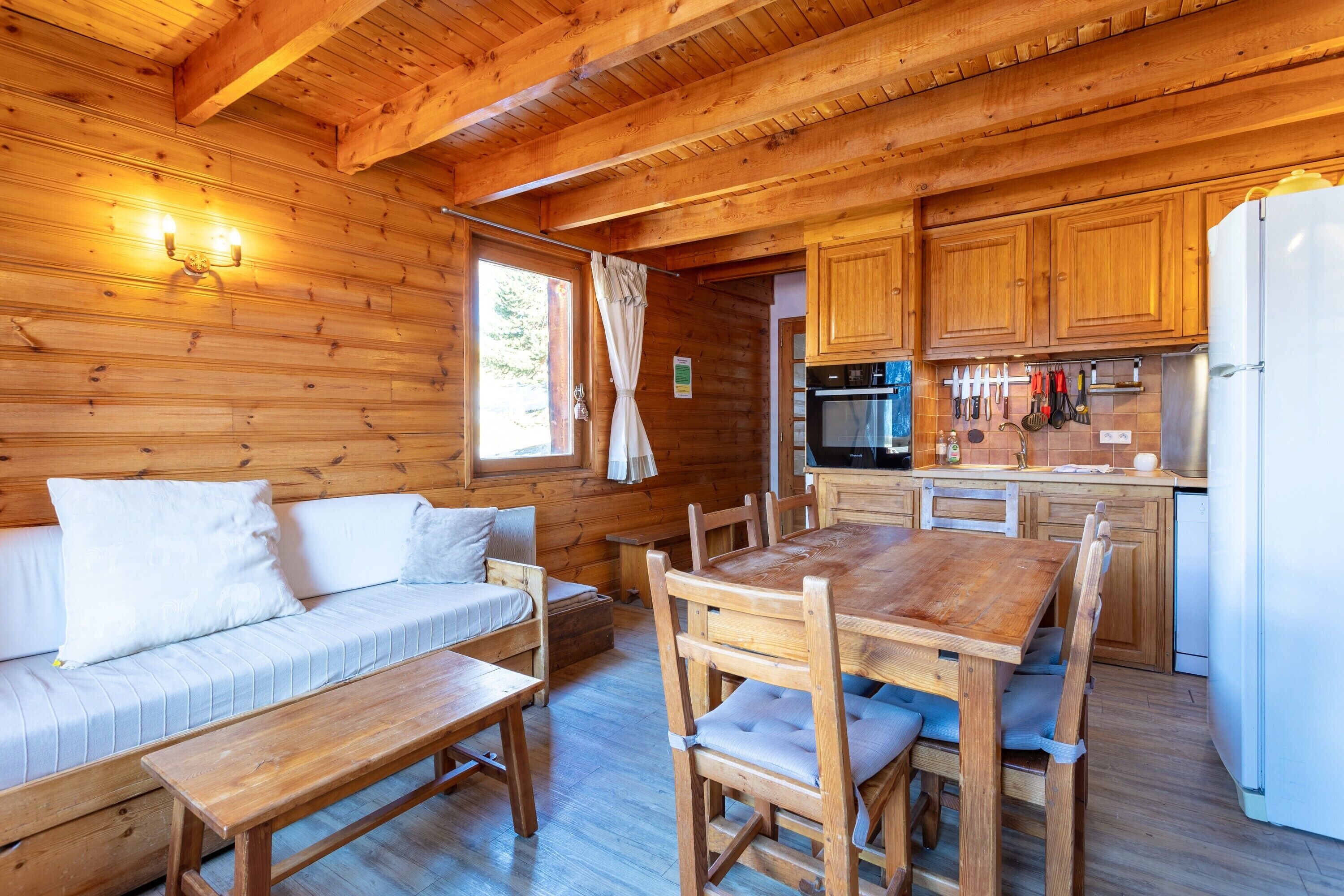 A wooden cabin interior with a sofa, dining table, chairs, wooden cabinets, a refrigerator, and a kitchen area.