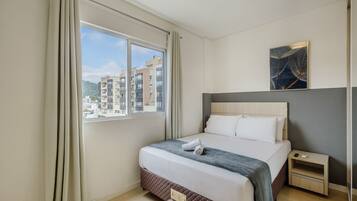 Quarto de hotel com uma cama grande, uma mesinha de cabeceira, uma janela com cortinas e vista para a cidade.