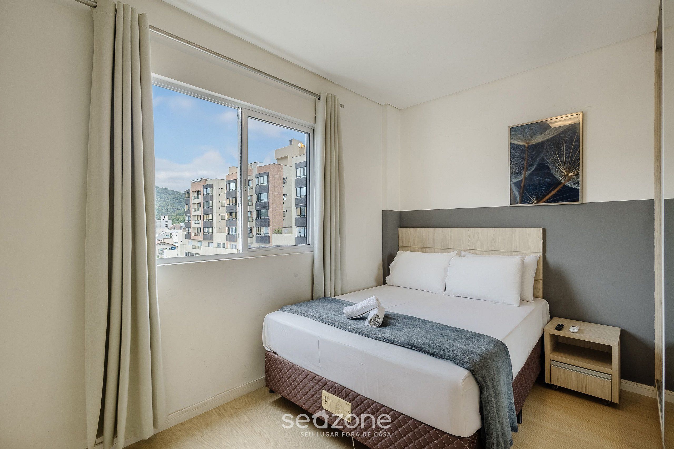 Quarto de hotel com uma cama grande, uma mesinha de cabeceira, uma janela com cortinas e vista para a cidade.