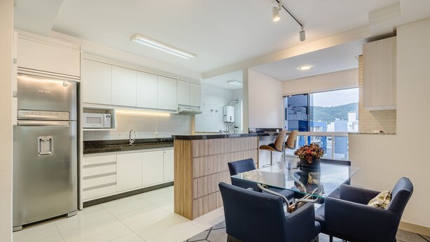 A modern kitchen with a stainless steel refrigerator, a microwave, and a water dispenser. There is a dining area with a glass table and blue chairs. - Apt w/ BBQ Garage 550m From Beach Zuc0703 (Balneário Camboriú)