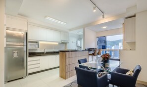 A modern kitchen with a stainless steel refrigerator, a microwave, and a water dispenser. There is a dining area with a glass table and blue chairs. - Apt w/ BBQ Garage 550m From Beach Zuc0703 (Balneário Camboriú)