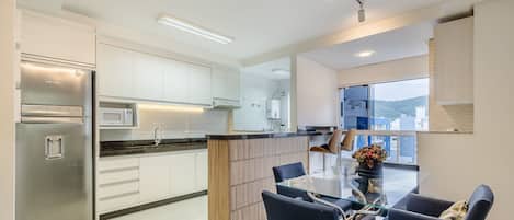 A modern kitchen with a stainless steel refrigerator, a microwave, and a water dispenser. There is a dining area with a glass table and blue chairs.