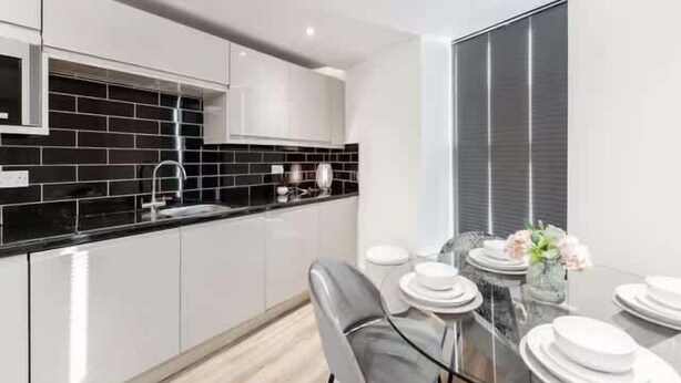 Una cocina moderna con armarios blancos, salpicadero negro y un comedor con mesa y sillas de vidrio.