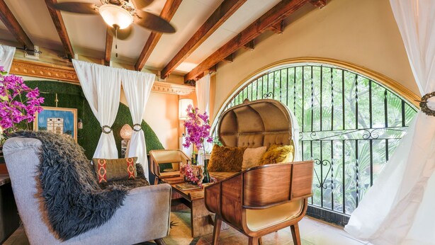 A cozy room with a wooden ceiling, a large window with a metal gate, a cushioned chair, and a table with a vase of flowers. - House of Mosaic Aruba - Unique Experience (Oranjestad)