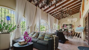 A spacious living room with a black sofa, a coffee table, and a chandelier. There are large windows with white curtains, a potted plant, and an easel with a painting. - House of Mosaic Aruba - Unique Experience (Oranjestad)