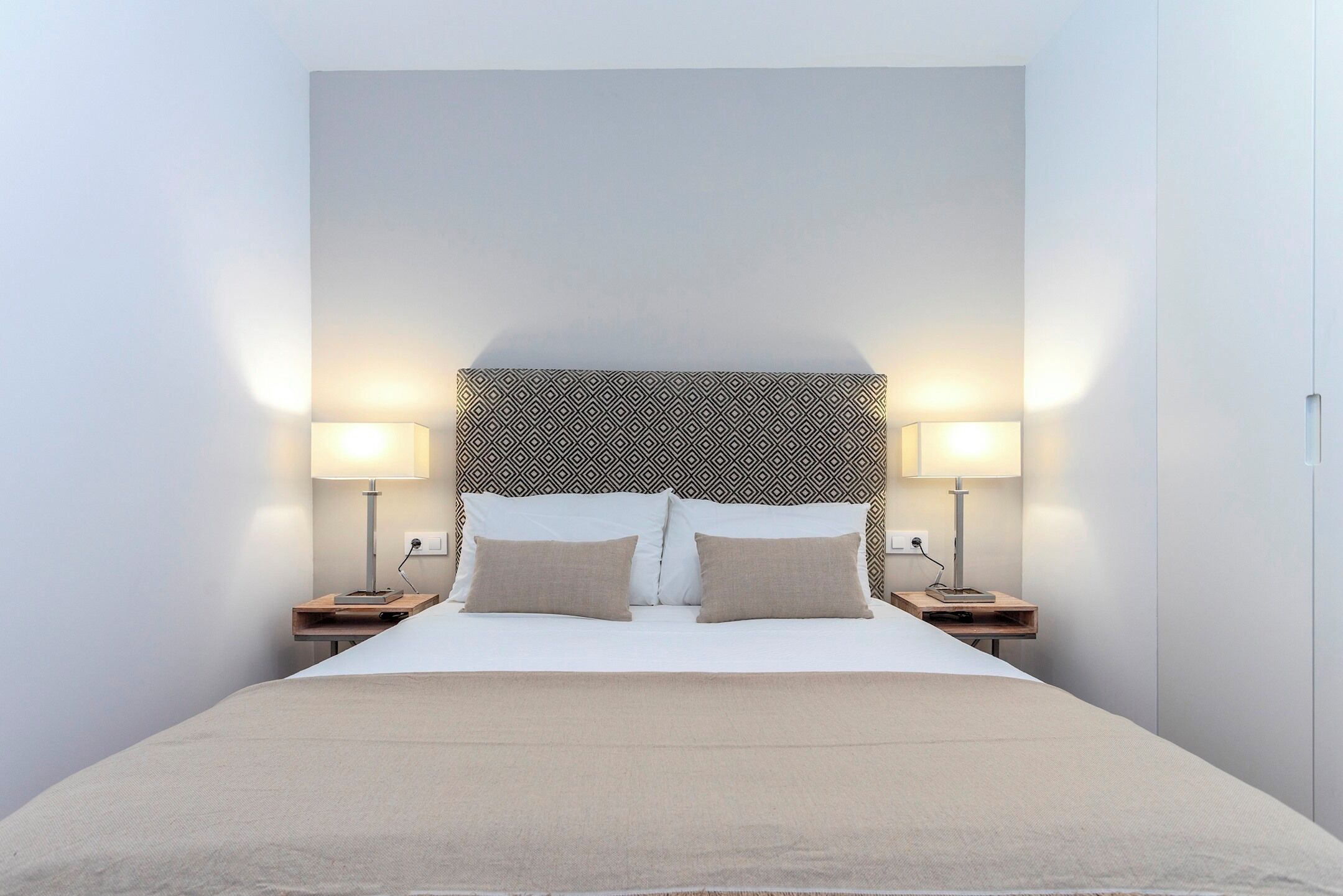 A neatly made bed with two bedside tables, each with a lamp, in a minimalist room with a patterned headboard.