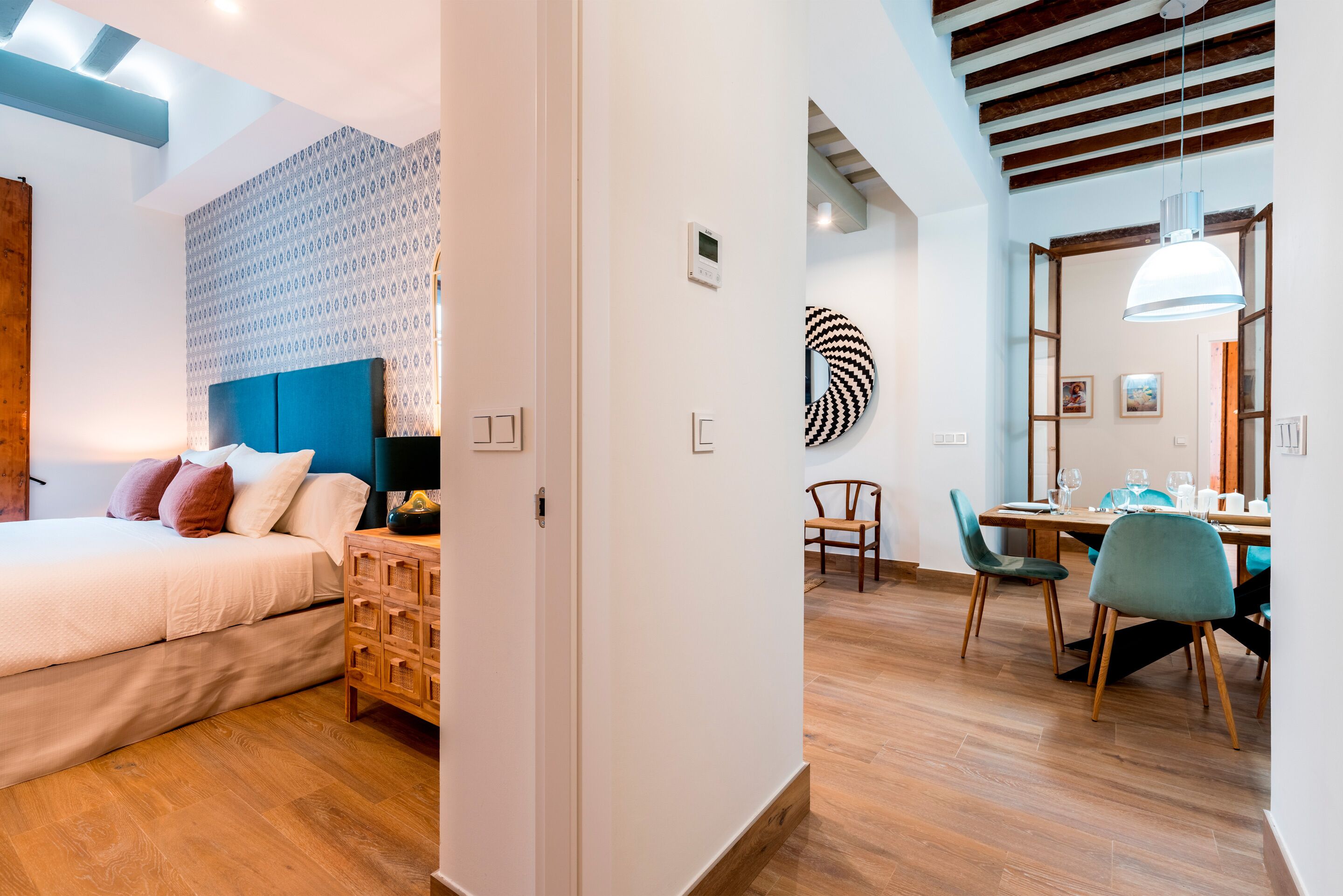 A modern hotel room with a bed, a wooden dresser, a dining area with a table and chairs, and a patterned wall.