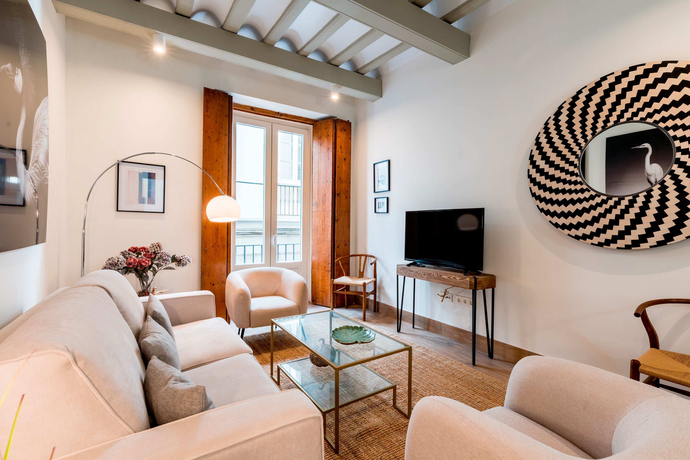 Una sala de estar con un sofá, sillones, una mesa de centro de vidrio, un televisor y un espejo decorativo.