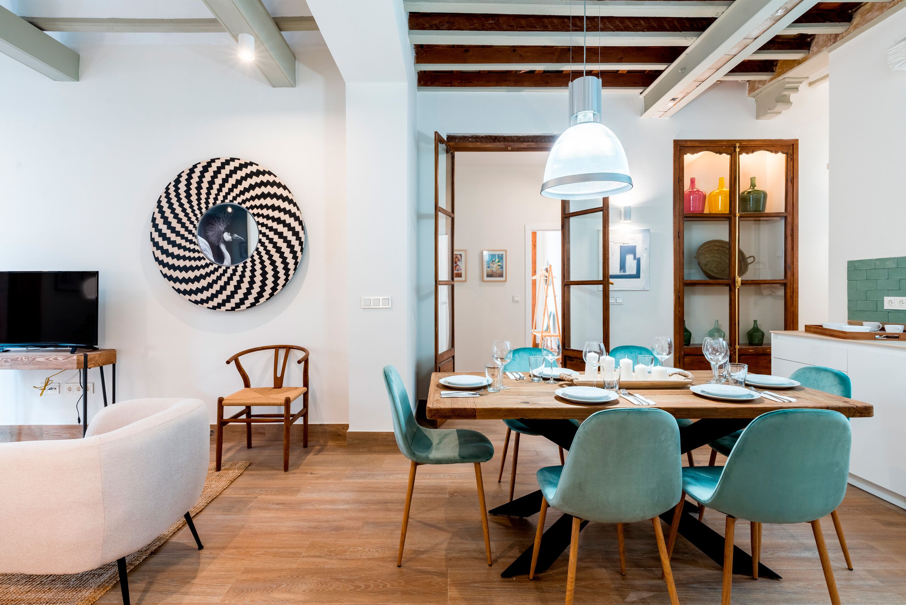 Un comedor con una mesa de madera dispuesta para cuatro personas, rodeada de sillas color turquesa. Un espejo grande cuelga en la pared sobre la mesa, y un televisor está montado en la pared de enfrente.